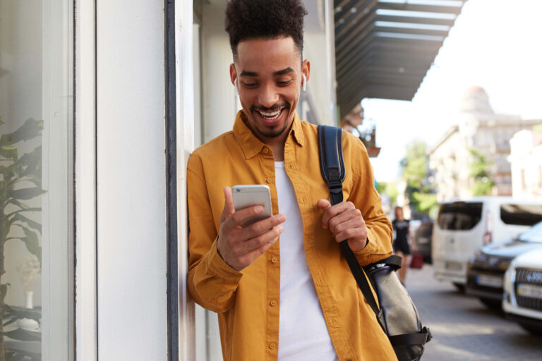 Young happy dark skinned man in yellow shirt, walking down the street and holds telephone, got a message with a funny video, looks joyful and broadly smiling.